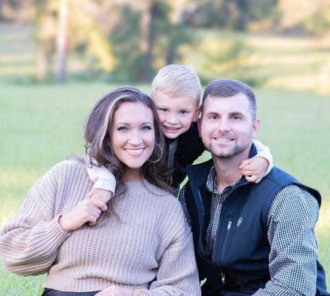 Family of three smiling outdoors with green field background. Woman in beige sweater, young blonde child, and man in navy vest with checkered shirt.