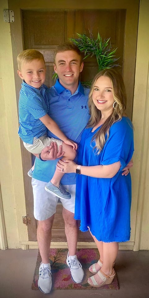 Family of three smiling indoors. Man in blue polo holding young boy in striped shirt, standing with woman in blue dress